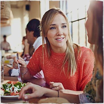 O femeie într-o cămașă portocalie stă cu un prieten în restaurant și mănâncă o salată.